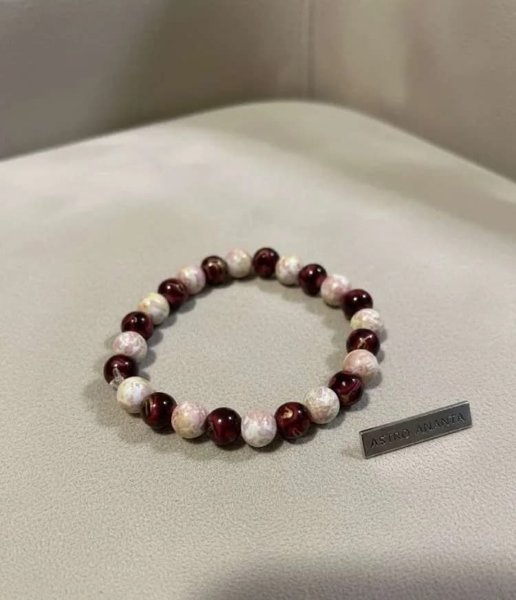 A crystal bead bracelet with alternating dark red Garnet and mottled pink Rhodonite beads on a neutral surface next to an 'Astro Ananta' metal tag.