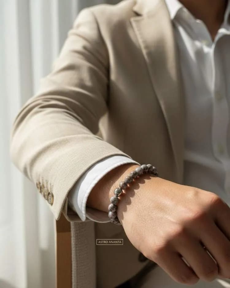 A man wearing a beige suit jacket and white shirt wears a grey and black beaded Larvikite bracelet on his wrist.