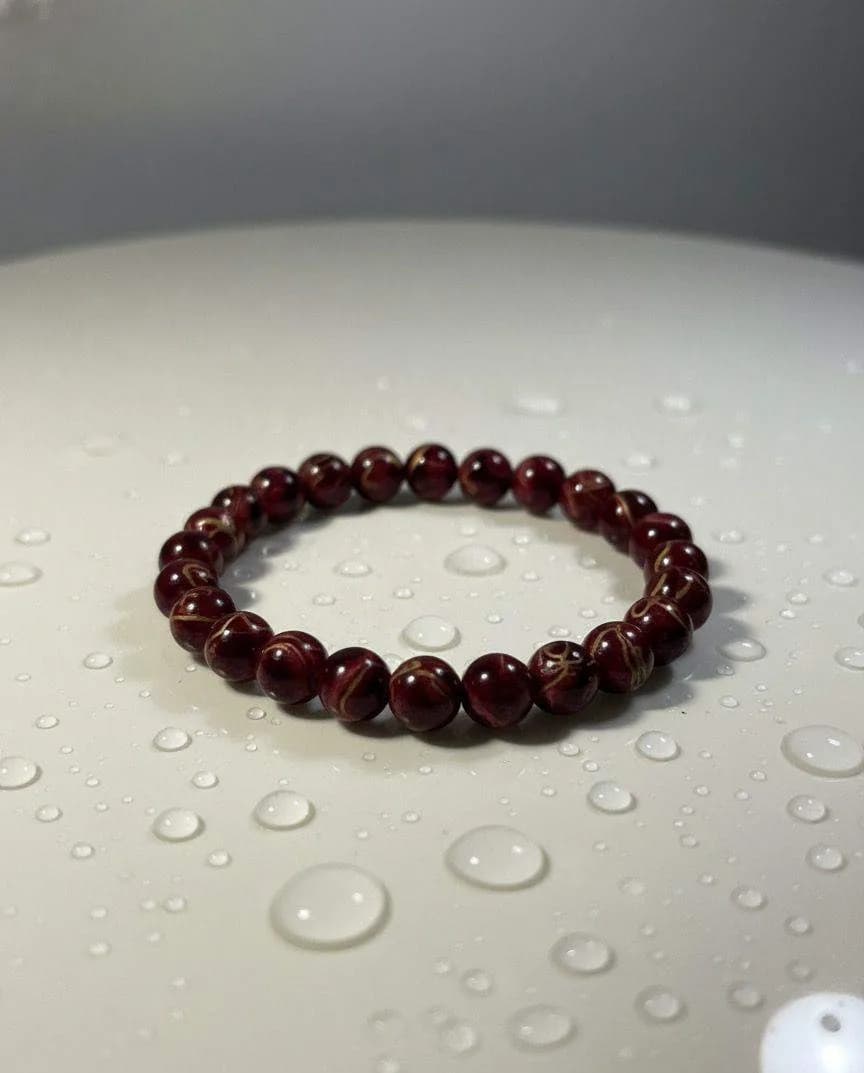 A Red Tiger's Eye beaded bracelet with deep red and subtle golden-brown bands, resting on a white surface with water droplets.