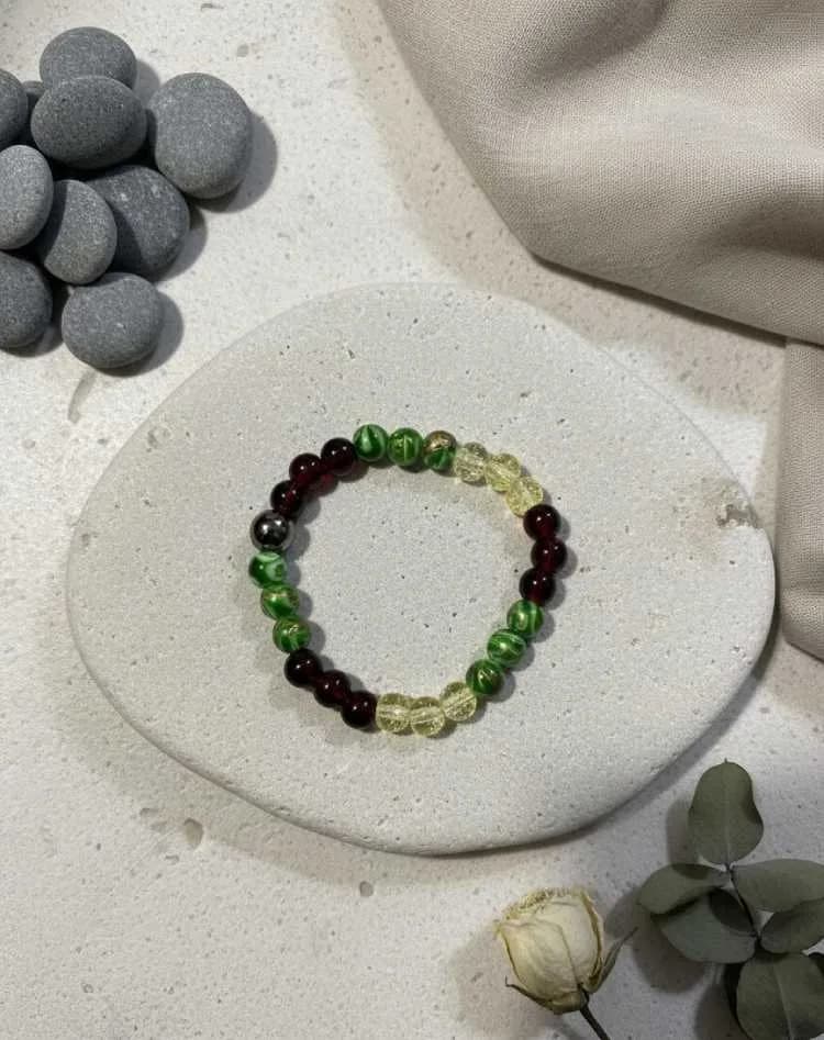 A crystal bracelet with yellow Citrine, green Green Aventurine, and dark red Garnet beads on a white stone coaster, next to grey pebbles.