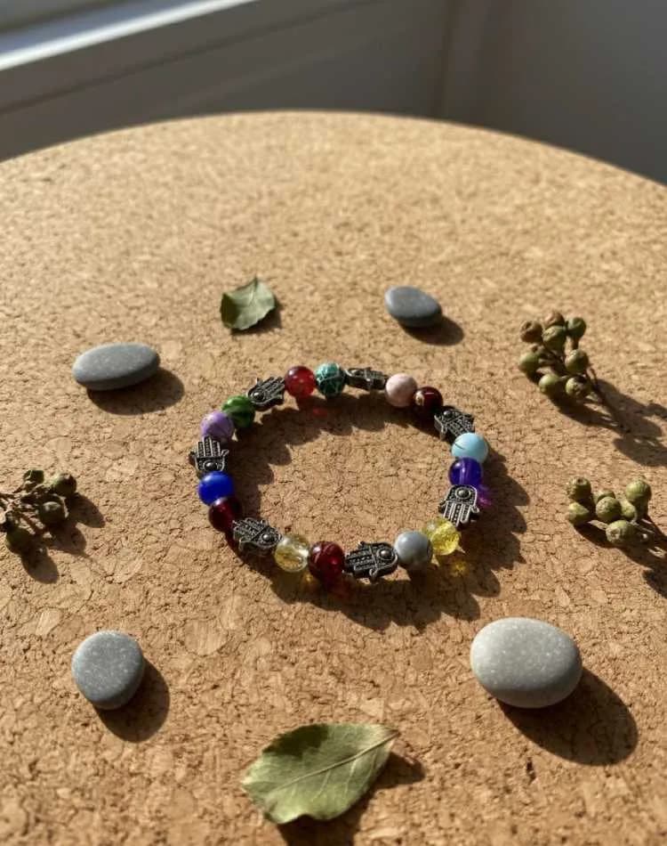 A 7-chakra bracelet with multi-colored beads and silver Hamsa Hand charms, arranged on a round cork board with small stones and leaves.