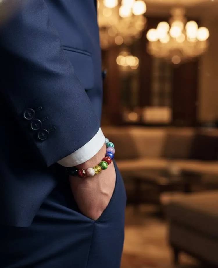 A man in a dark blue suit wearing a polished, multi-colored 7 Chakra beaded bracelet on his wrist in a luxurious setting with a chandelier visible.