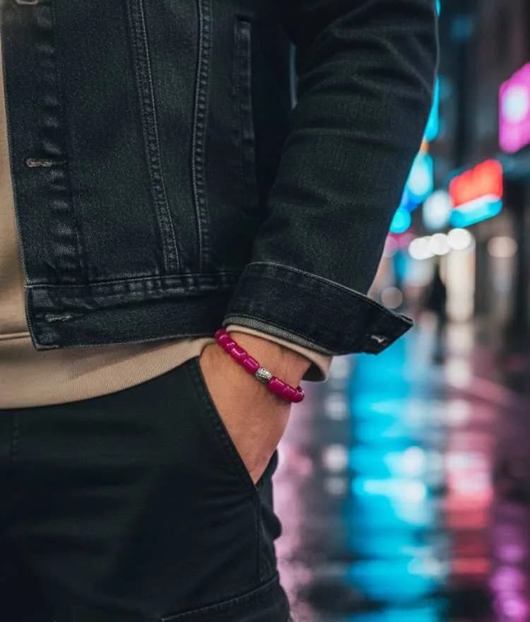 A person wearing a bright magenta/hot pink beaded bracelet with a silver metal spacer, standing on a wet street at night with neon lights reflecting on the ground.
