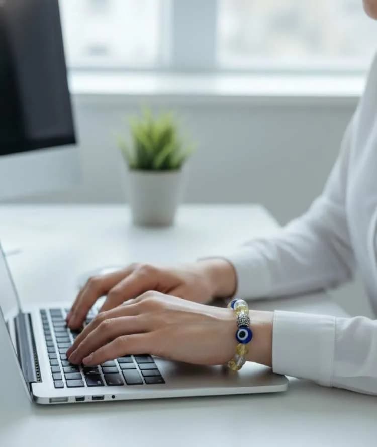 A woman typing on a laptop, wearing a yellow Citrine beaded bracelet with a large blue and yellow Evil Eye charm on her wrist.