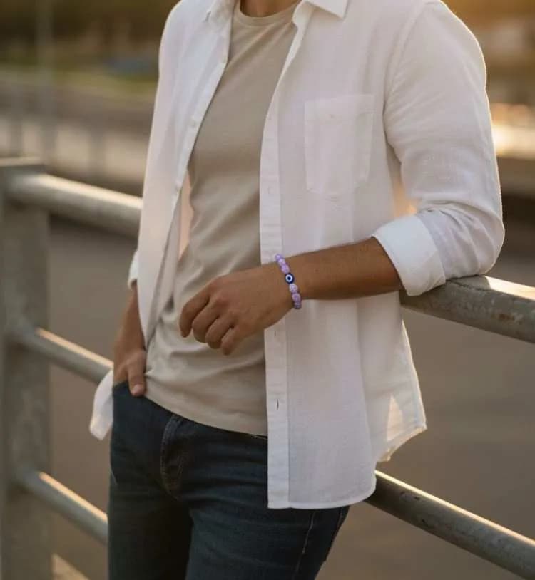 A man leaning on a railing at sunset, wearing a light purple Lavender Quartz beaded bracelet with a large blue Evil Eye charm.
