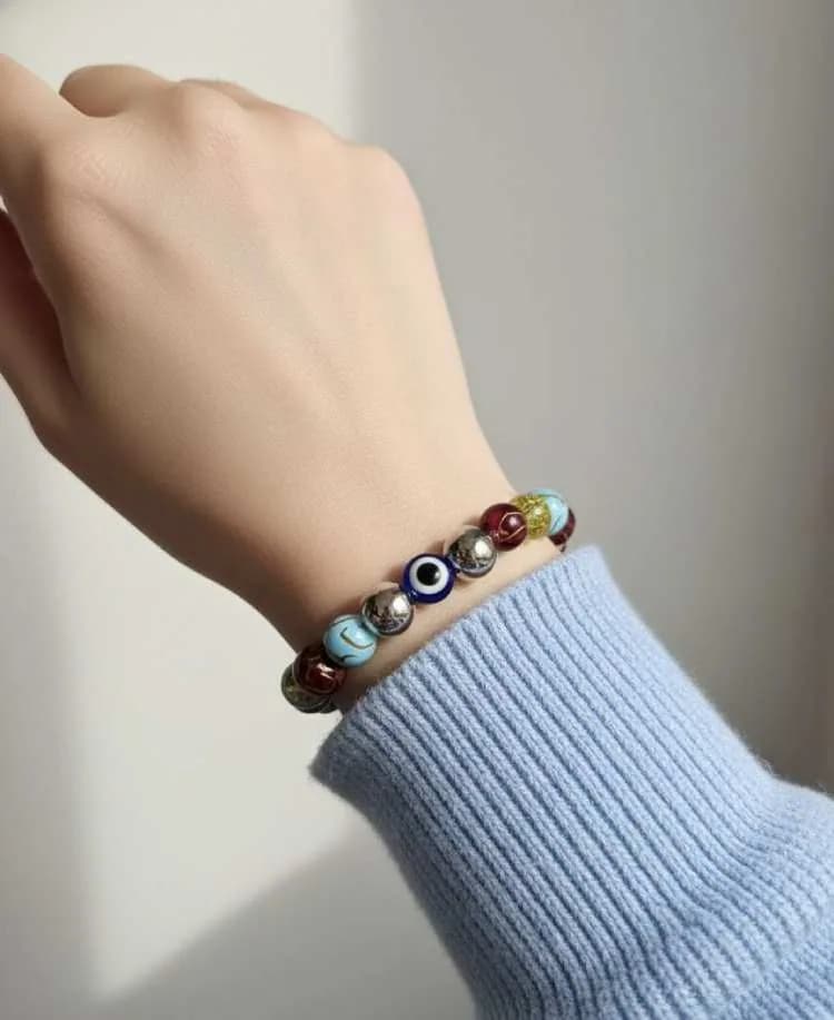 A person wearing a blue sweater wears a multi-colored beaded bracelet with a prominent blue Evil Eye charm and silver spacers on their wrist.A person wearing a blue sweater wears a multi-colored beaded bracelet with a prominent blue Evil Eye charm and sil