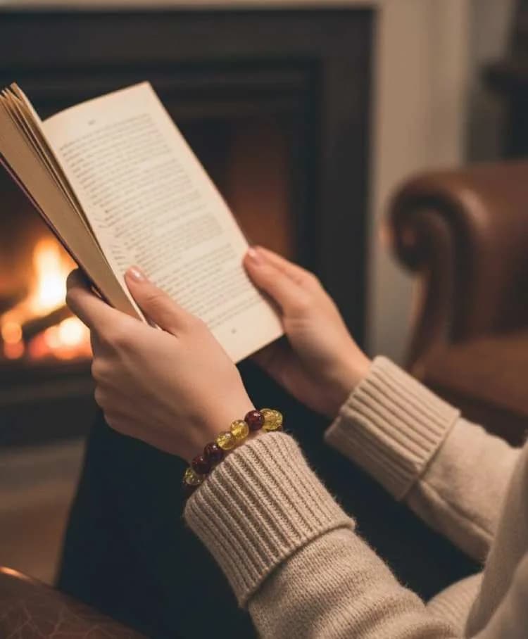A person sitting by a fireplace, reading a book, wearing an alternating red Garnet and yellow Citrine beaded bracelet on their wrist.