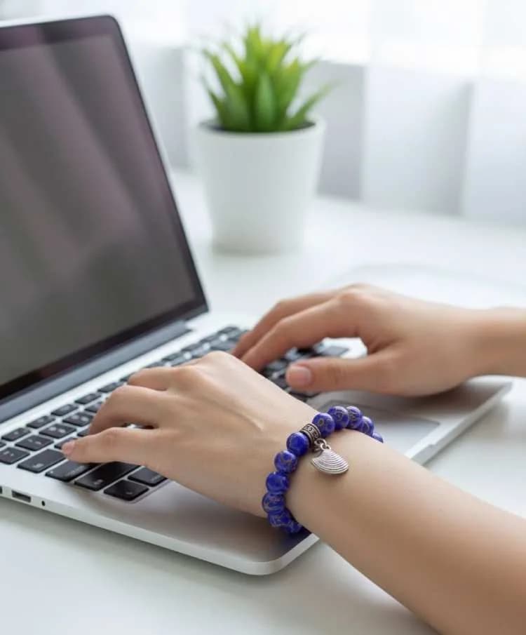 A person typing on a laptop, wearing a vibrant blue Imperial Jasper beaded bracelet with a silver shell charm on their wrist.