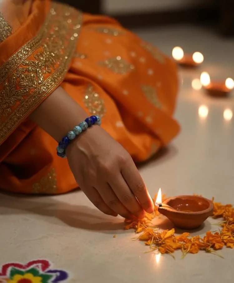 A person in an orange sari performing a ritual (Pooja/Diwali), lighting a diya while wearing a dual blue patterned beaded bracelet on their wrist.