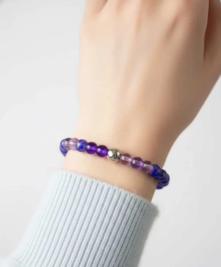 A person wearing a light blue sweater wears a dual-toned purple Amethyst and patterned beaded bracelet with a silver spacer on their wrist.