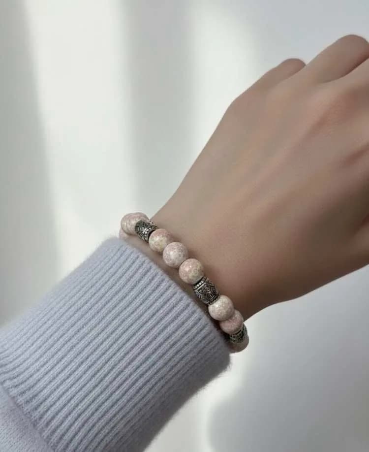 A person wearing a light blue sweater wears a pale pink Rhodonite beaded bracelet with antique silver patterned spacers on their wrist.