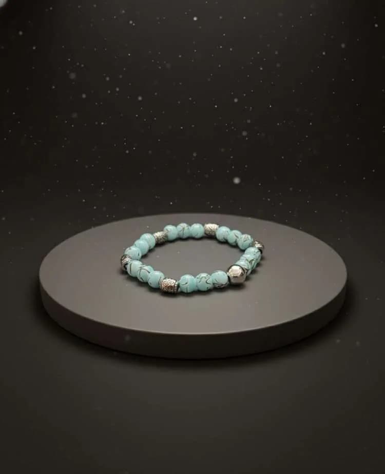 A light blue Turquoise beaded bracelet with black matrix and antique silver spacers, displayed on a grey circular pedestal in a dark setting.