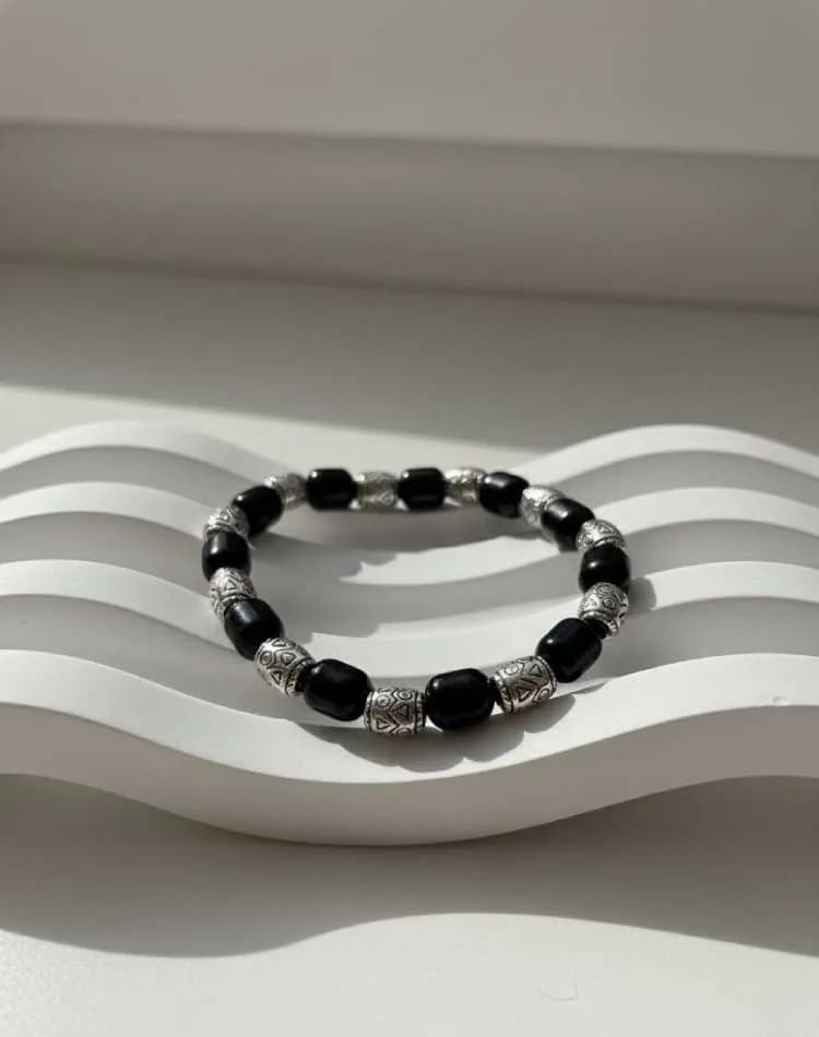 A Black Onyx beaded bracelet with antique silver patterned cylinder spacers, resting on a wavy white textured surface in sunlight.