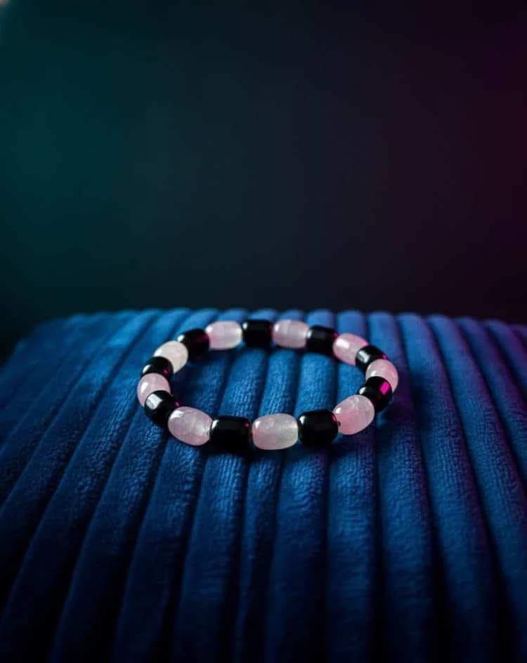 A bracelet with alternating pink Rose Quartz and black Onyx barrel-shaped beads, resting on a deep blue pleated velvet cushion.