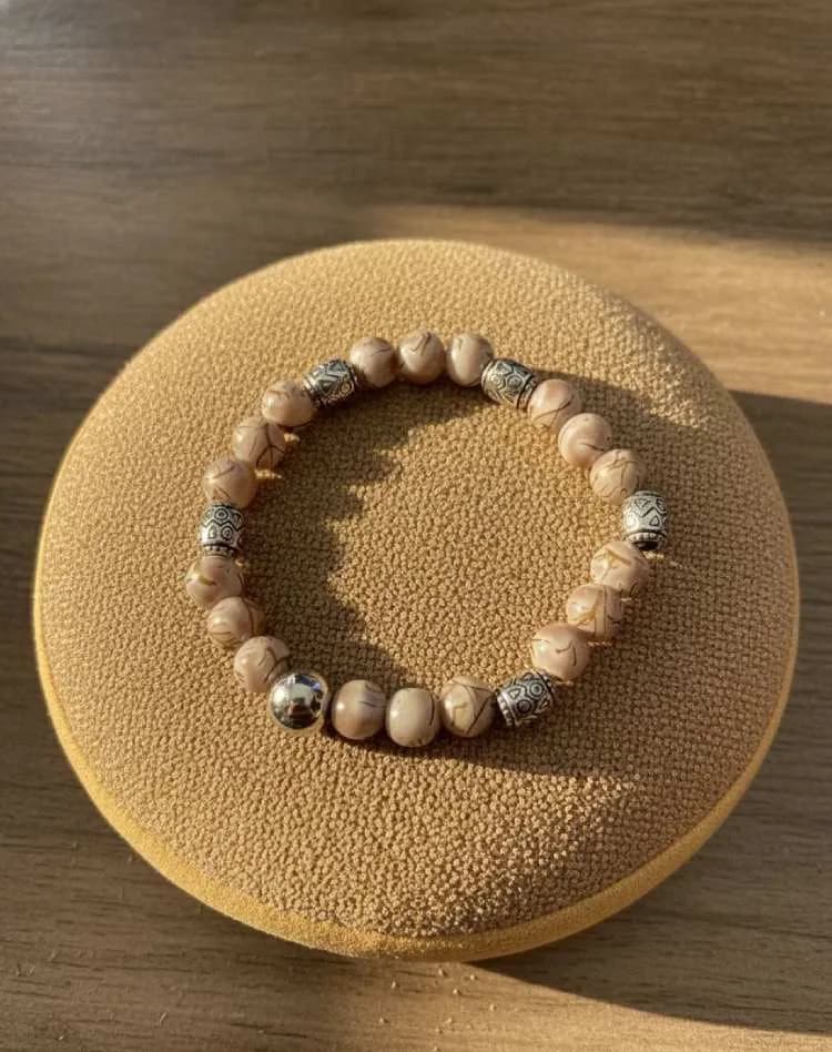A beige/tan patterned Picture Jasper beaded bracelet with silver ornate spacers, resting on a round, suede-like cushion.
