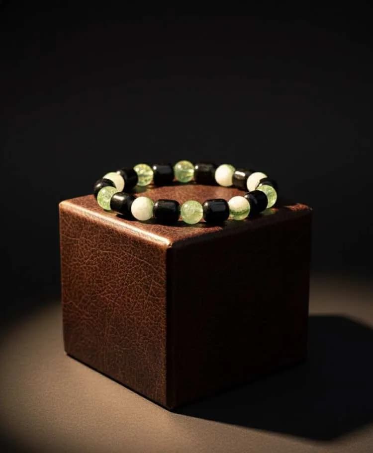 A bracelet with green Aventurine, black Onyx, and white Jade beads with silver spacers, displayed on a small brown leather cube.