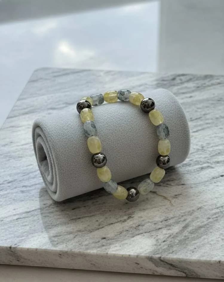 A bracelet with alternating pale yellow Lemon Quartz and grey Smoky Quartz barrel-shaped beads, with metallic Hematite spacers, resting on a grey velvet roll on a marble surface.