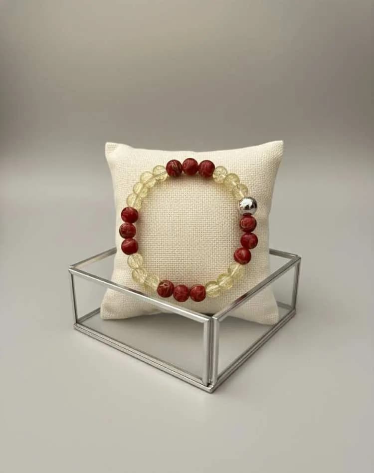 A bracelet with alternating yellow Citrine and patterned red Jasper beads, with a silver spacer, resting on a small white cushion on a glass display box.