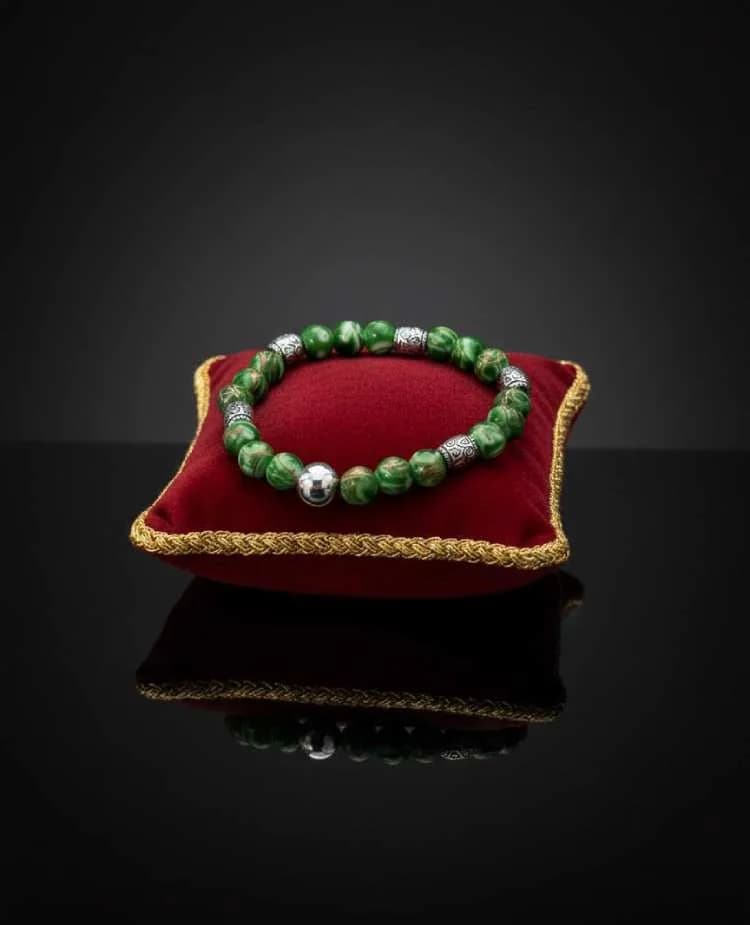 A mottled green Imperial Jasper beaded bracelet with silver ornate spacers, displayed on a small red velvet cushion.