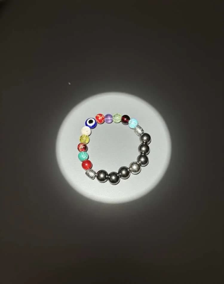 A bracelet combining multi-colored 7 Chakra beads, an Evil Eye charm, and dark metallic Hematite beads, illuminated by a spotlight on a dark background.
