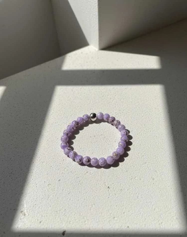 A light purple patterned Lilac Jasper beaded bracelet with a silver spacer, resting on a white textured surface in sunlight.